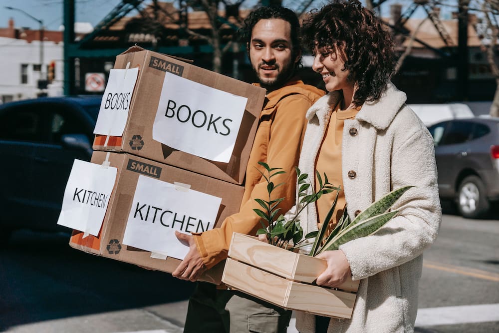 A many carries labeled boxes and a woman carries a plant as they are moving in together. Photo by Blue Bird