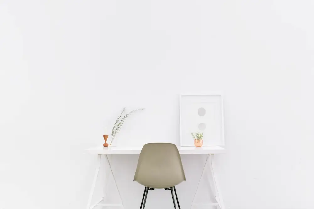 white desk in a minimalist white room with a white picture frame, one plant and a olive-colored desk chair